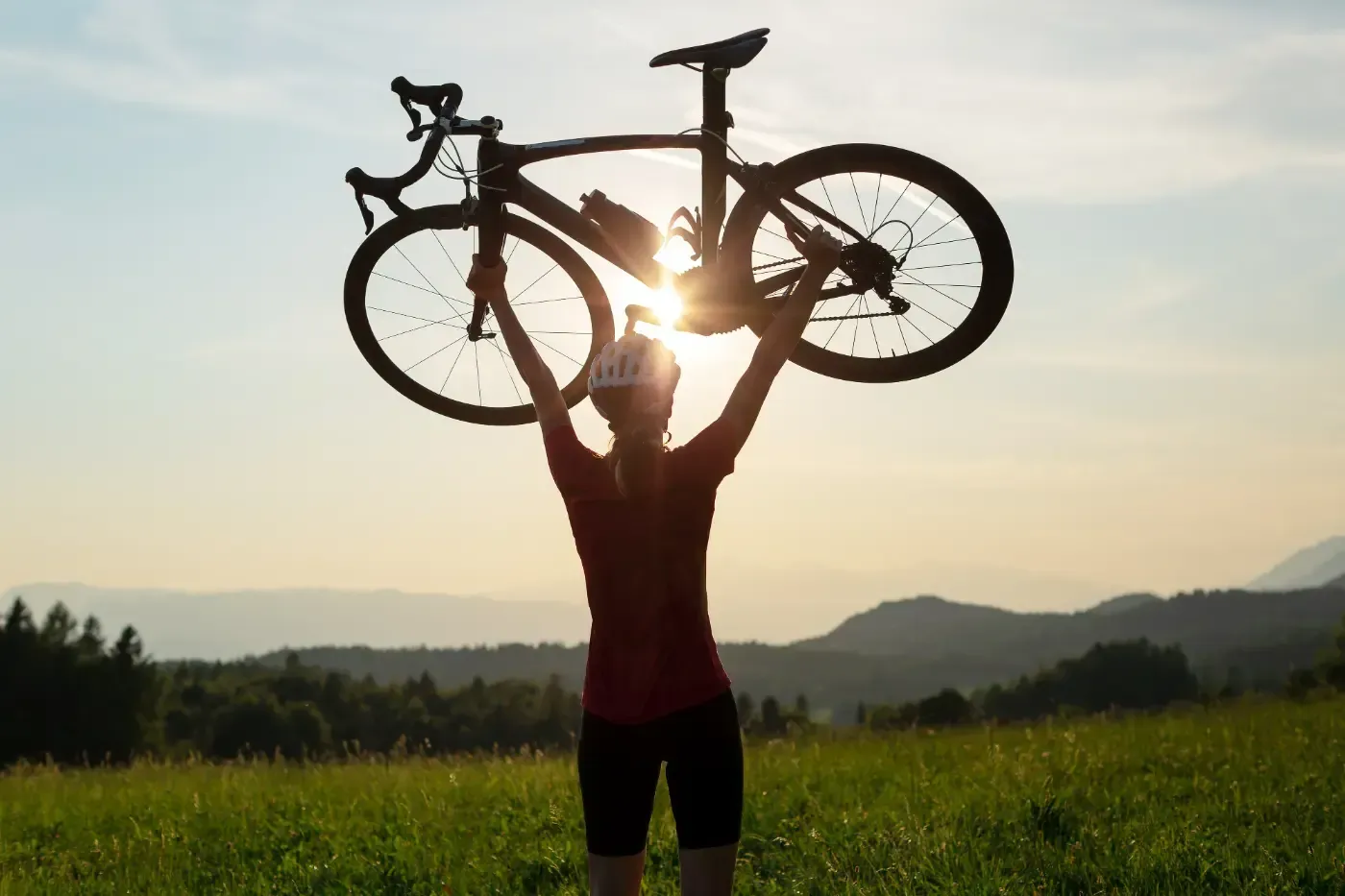 Ein Radfahrer steht bei Sonnenuntergang auf einer grasbewachsenen Wiese und hebt sein Fahrrad triumphierend gegen das Licht über den Kopf.