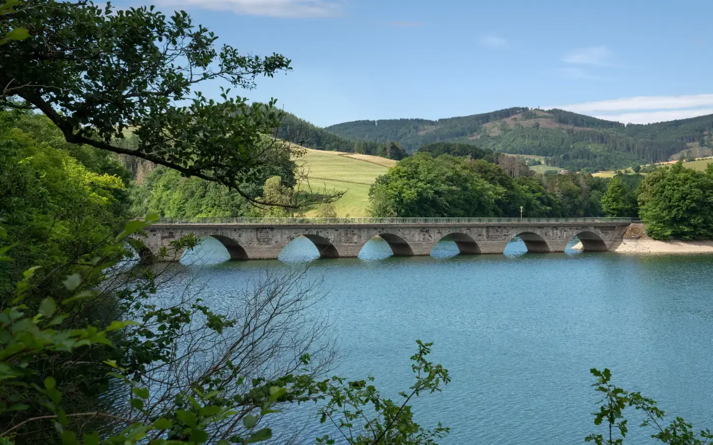 Eine Steinbrücke mit mehreren Bögen überspannt einen ruhigen blauen Stausee, der von üppig grünen Hügeln unter strahlend blauem Himmel umgeben ist.