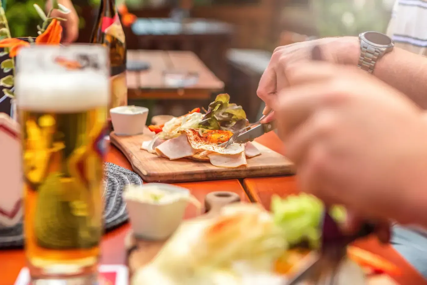Nahaufnahme einer Person, die an einem Gartentisch auf einem Holzbrett Essen schneidet; im Vordergrund steht ein Glas Bier.
