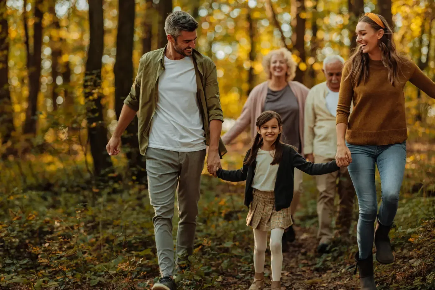 Eine Familie mehrerer Generationen spaziert Hand in Hand durch einen sonnenbeschienenen Herbstwald.