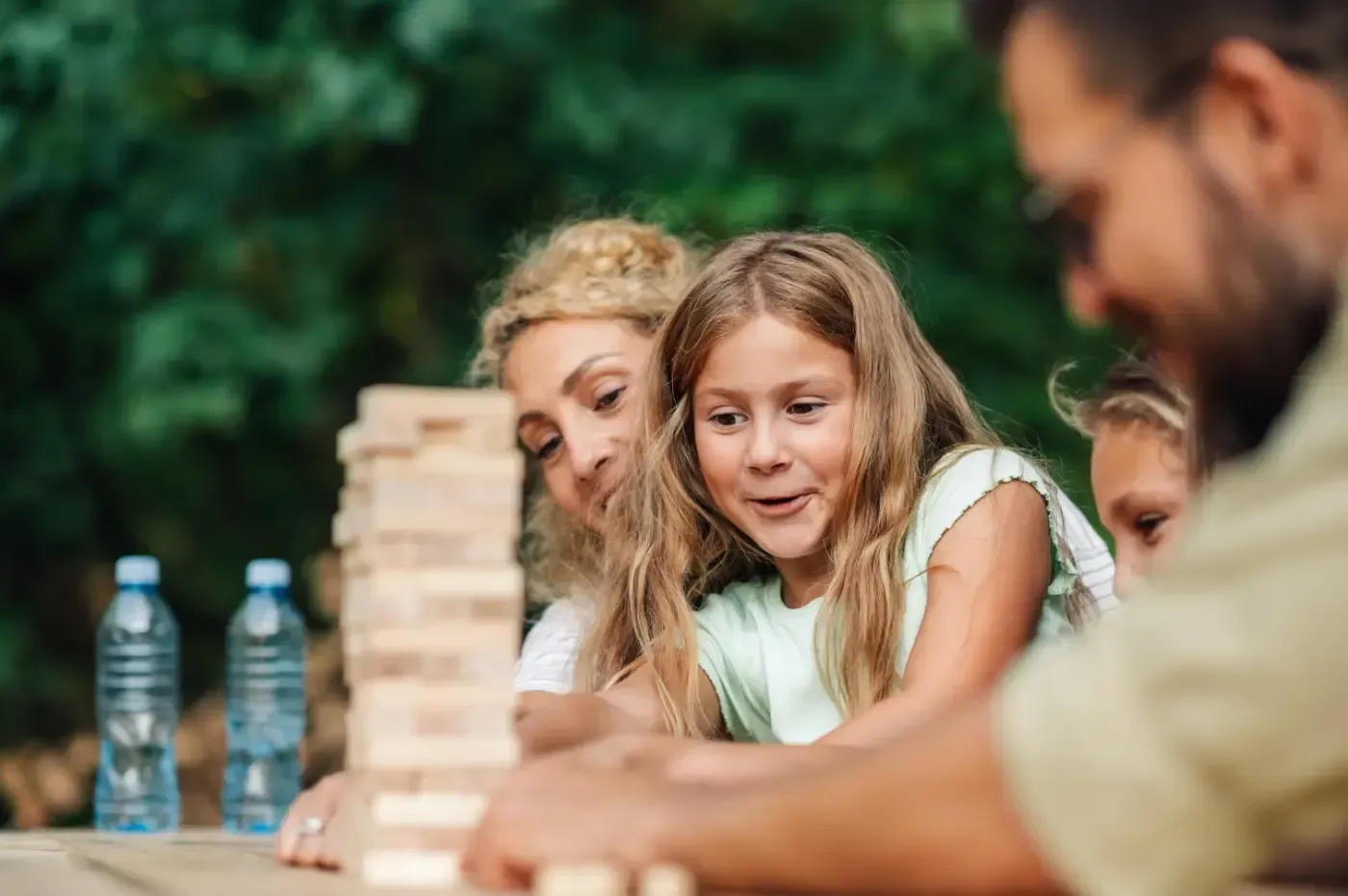 Eine Familie spielt im Freien Jenga an einem Holztisch; im Hintergrund sind Plastikwasserflaschen zu sehen.