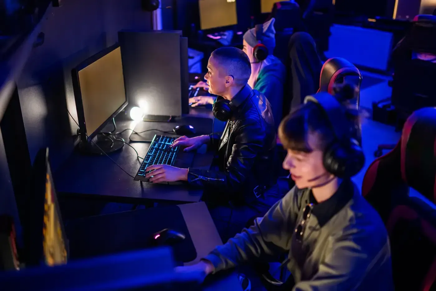 Menschen sitzen in einer schwach beleuchteten Gaming-Lounge, tragen Headsets und benutzen Computer mit leuchtenden Tastaturen.