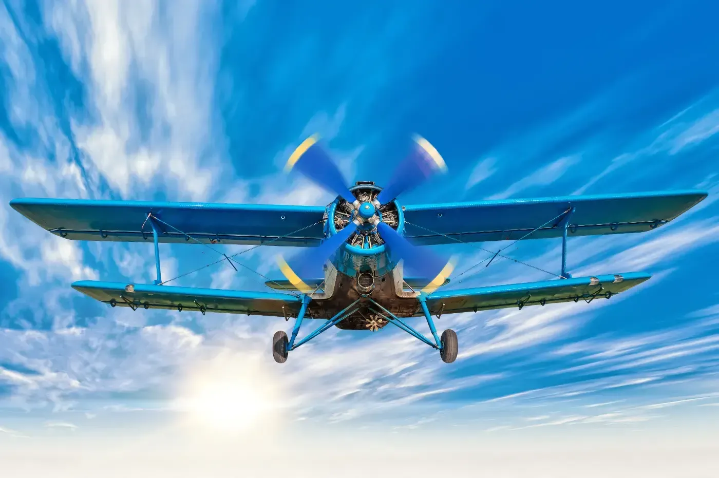 Ein blauer Doppeldecker mit sich drehendem Propeller fliegt vor einem hellen, sonnigen Himmel mit leichten Wolken.