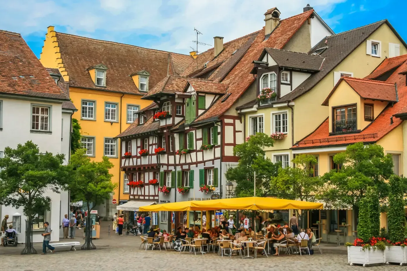 Menschen speisen an Tischen im Freien unter gelben Sonnenschirmen auf einem Platz, der von historischen Fachwerkhäusern umgeben ist.