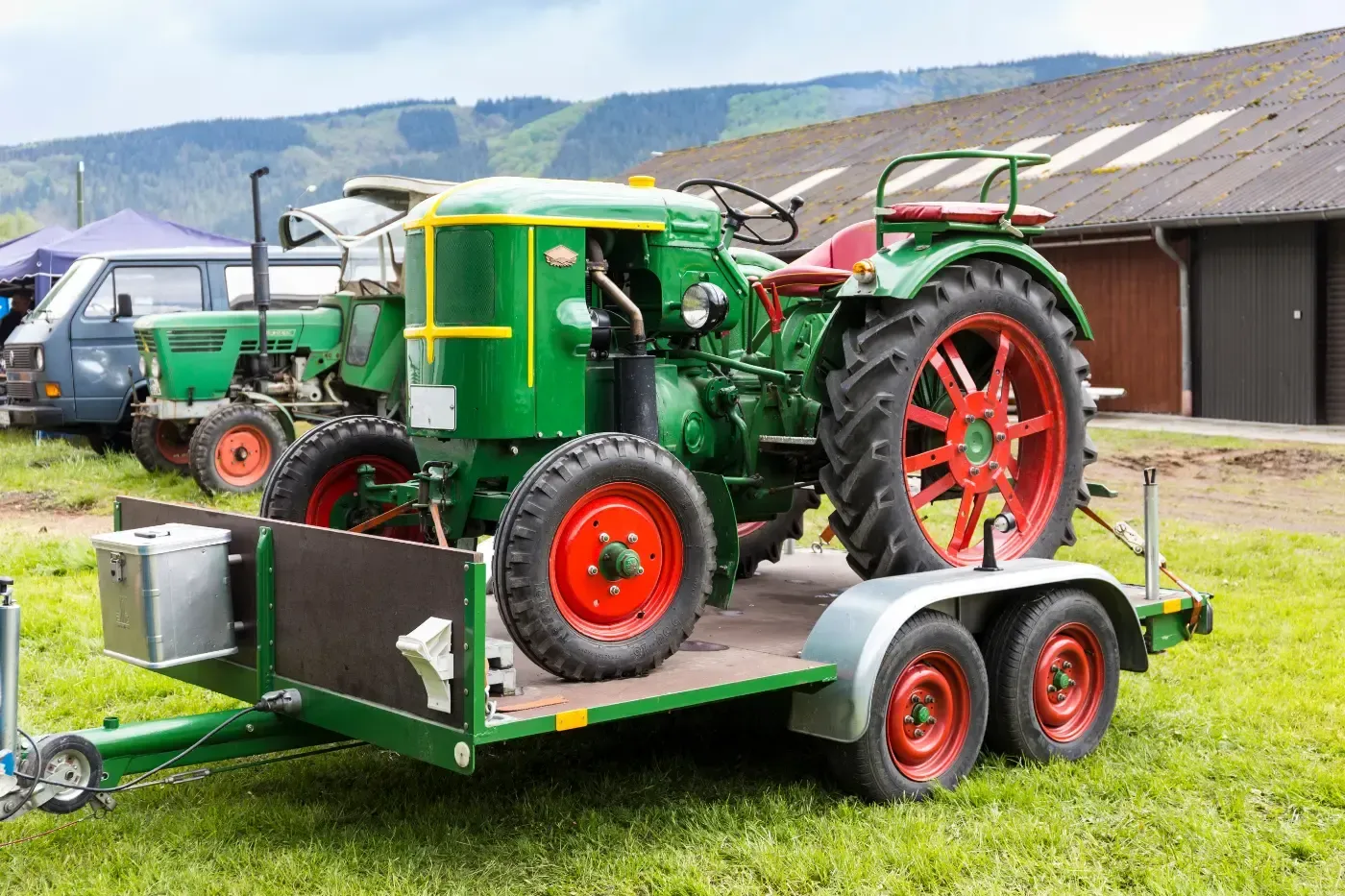 Ein grüner Oldtimer-Traktor mit roten Rädern, der auf einem Tieflader auf einem grasbewachsenen Freigelände gesichert ist.