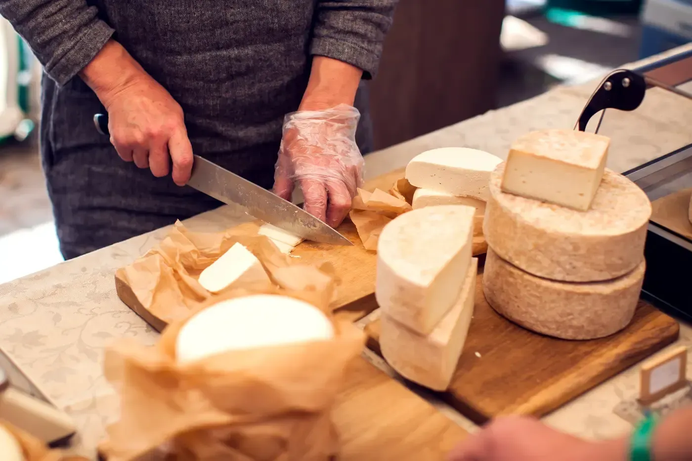 Eine Person in einem grauen Pullover und mit einem Plastikhandschuh schneidet Käse auf einem Holzbrett an einem Marktstand.