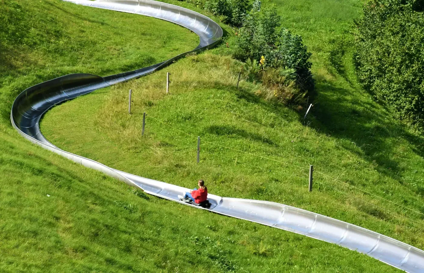 Eine Person in einem roten Hemd fährt mit einer Sommerrodelbahn eine gewundene Metallbahn hinunter, die in einen grasbewachsenen grünen Hang gebaut ist.
