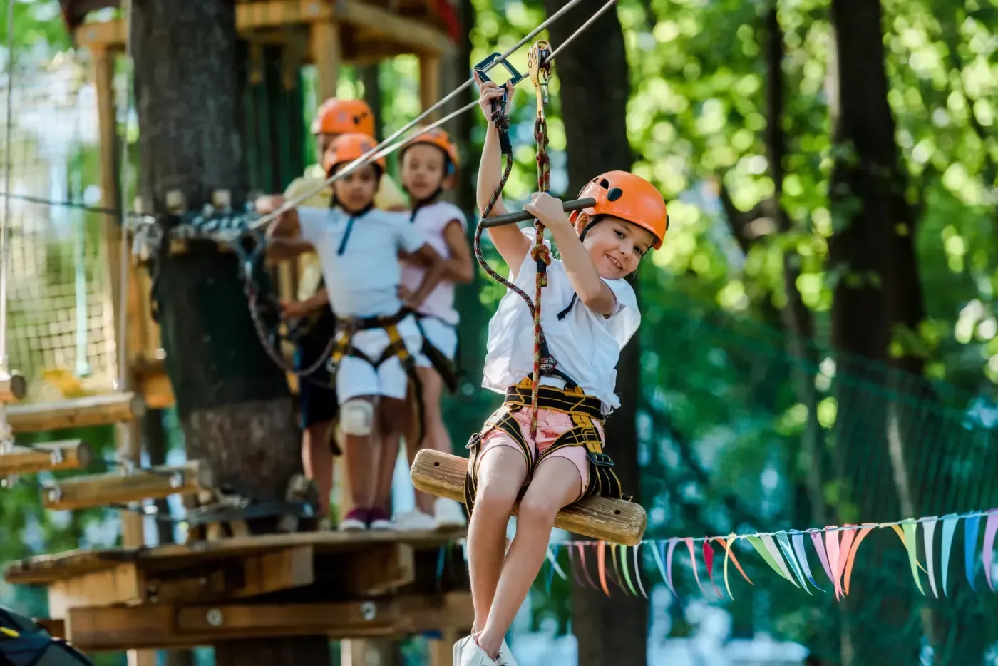 Ein Kind mit Helm und Gurtzeug an einer Seilrutsche, während andere Kinder auf einer Holzplattform in einer Waldlandschaft warten.