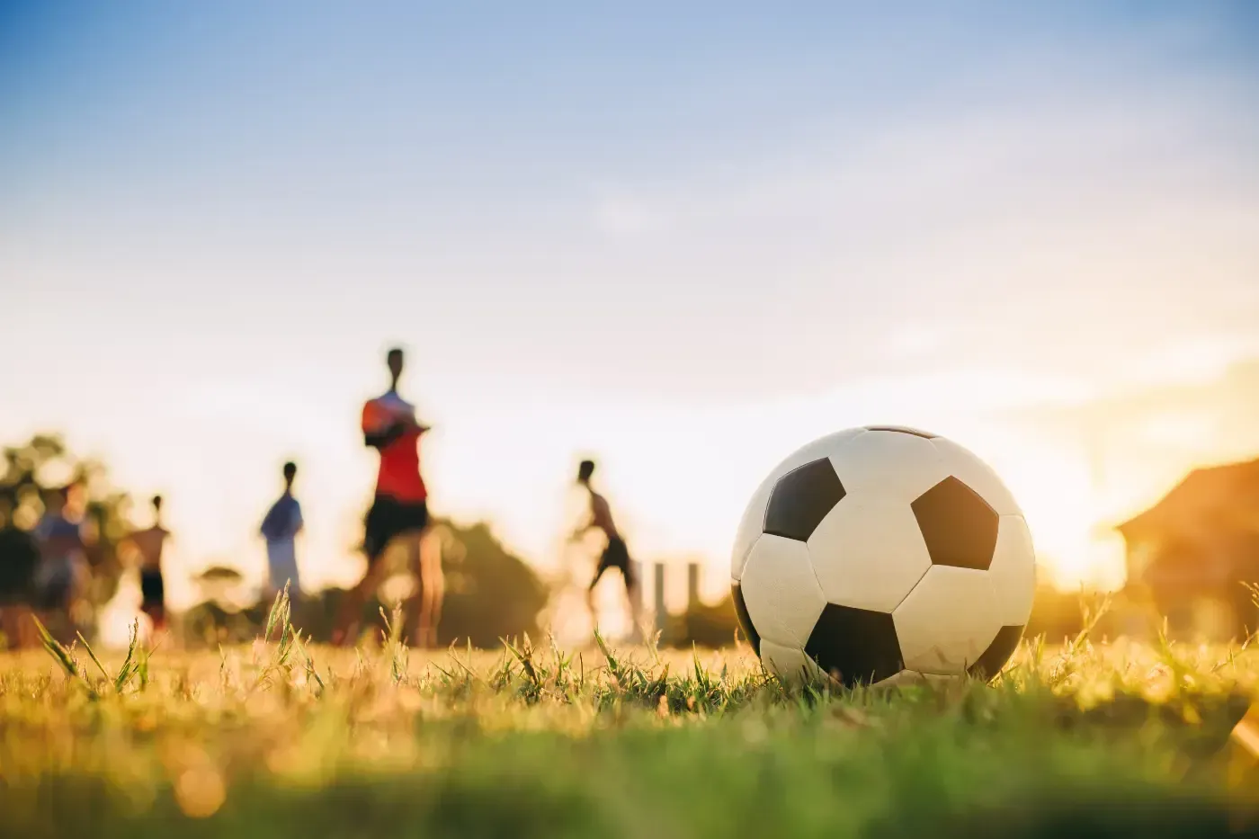 Im Vordergrund liegt ein Fußball auf einer Rasenfläche, im Hintergrund spielen verschwommene Gestalten bei Sonnenuntergang.