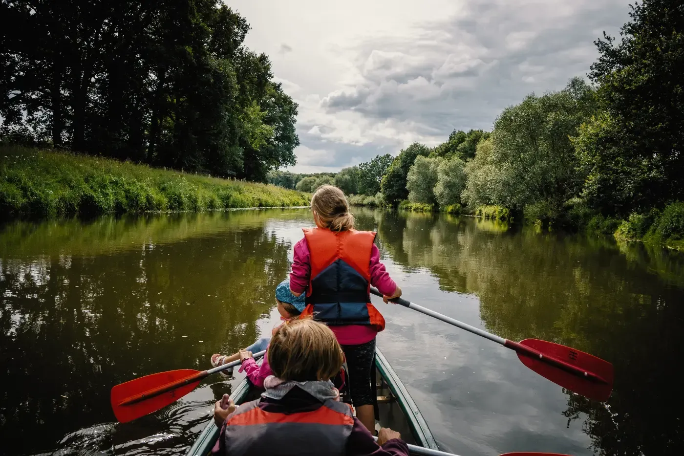 Drei Personen in Schwimmwesten paddeln in einem Kanu einen ruhigen, von Bäumen gesäumten Fluss unter bewölktem Himmel hinunter.