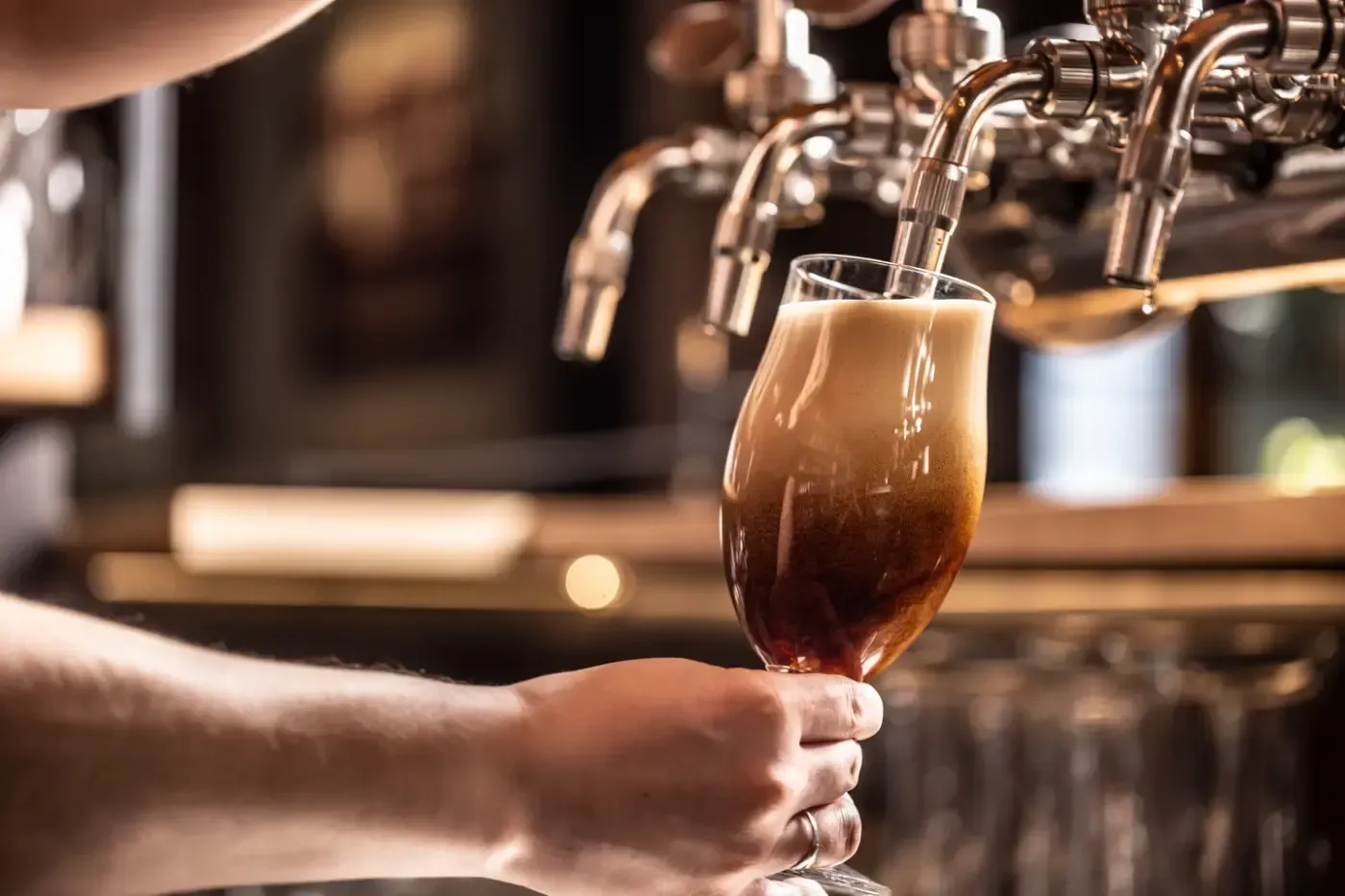 Eine Person zapft in einer schwach beleuchteten Bar ein dunkles, schaumiges Bier aus einem Metallhahn in ein Glas.