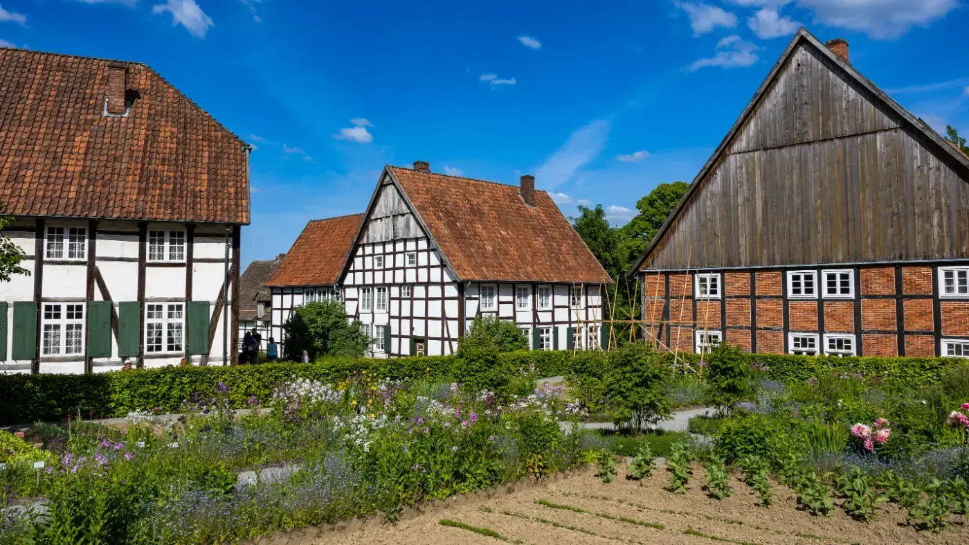 Drei traditionelle Fachwerkhäuser mit roten Ziegeldächern stehen hinter einem üppigen Garten unter strahlend blauem Himmel.