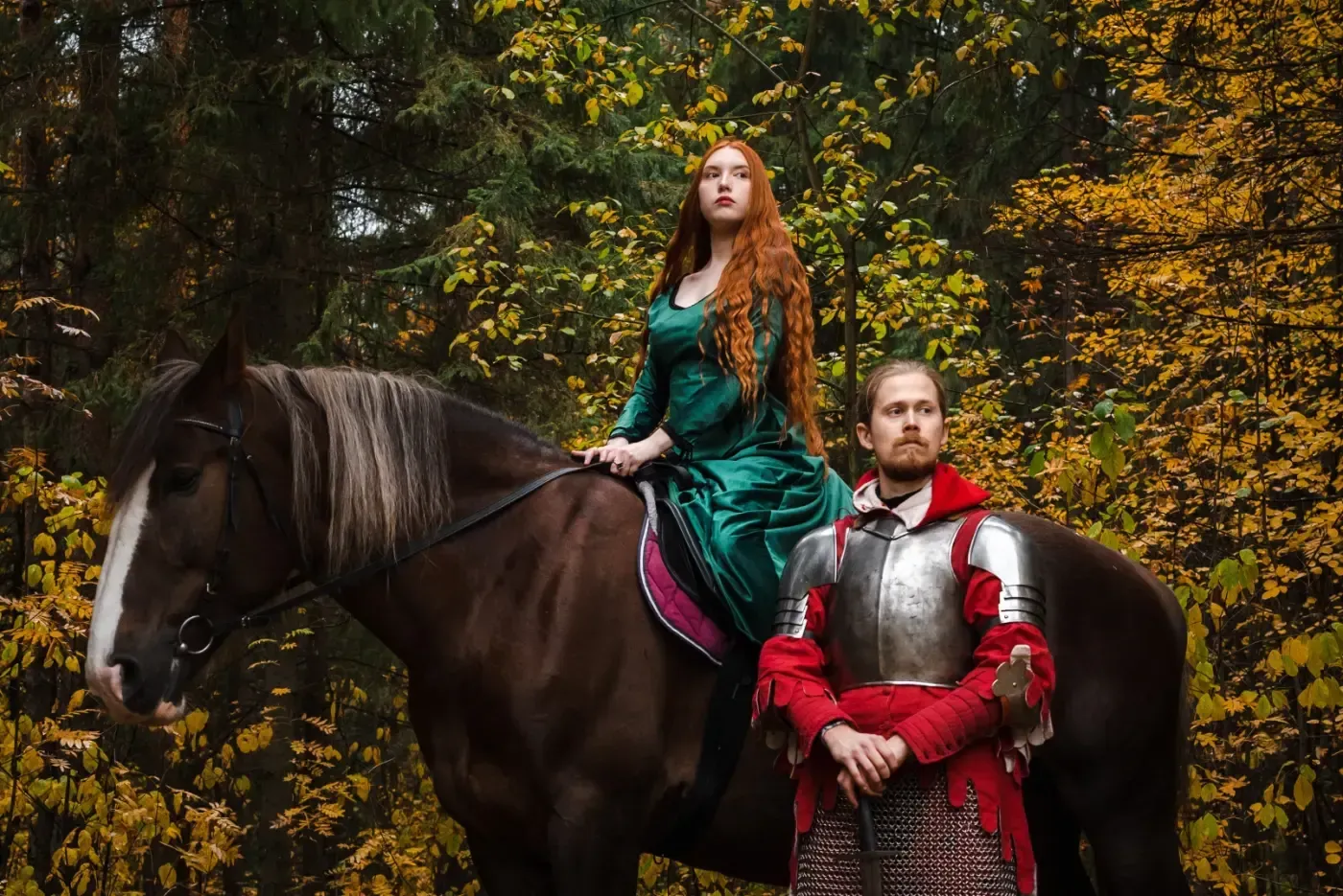 In einem Herbstwald steht eine Person in mittelalterlicher Rüstung neben einem Pferd, das eine Person in einem grünen Samtkleid trägt.