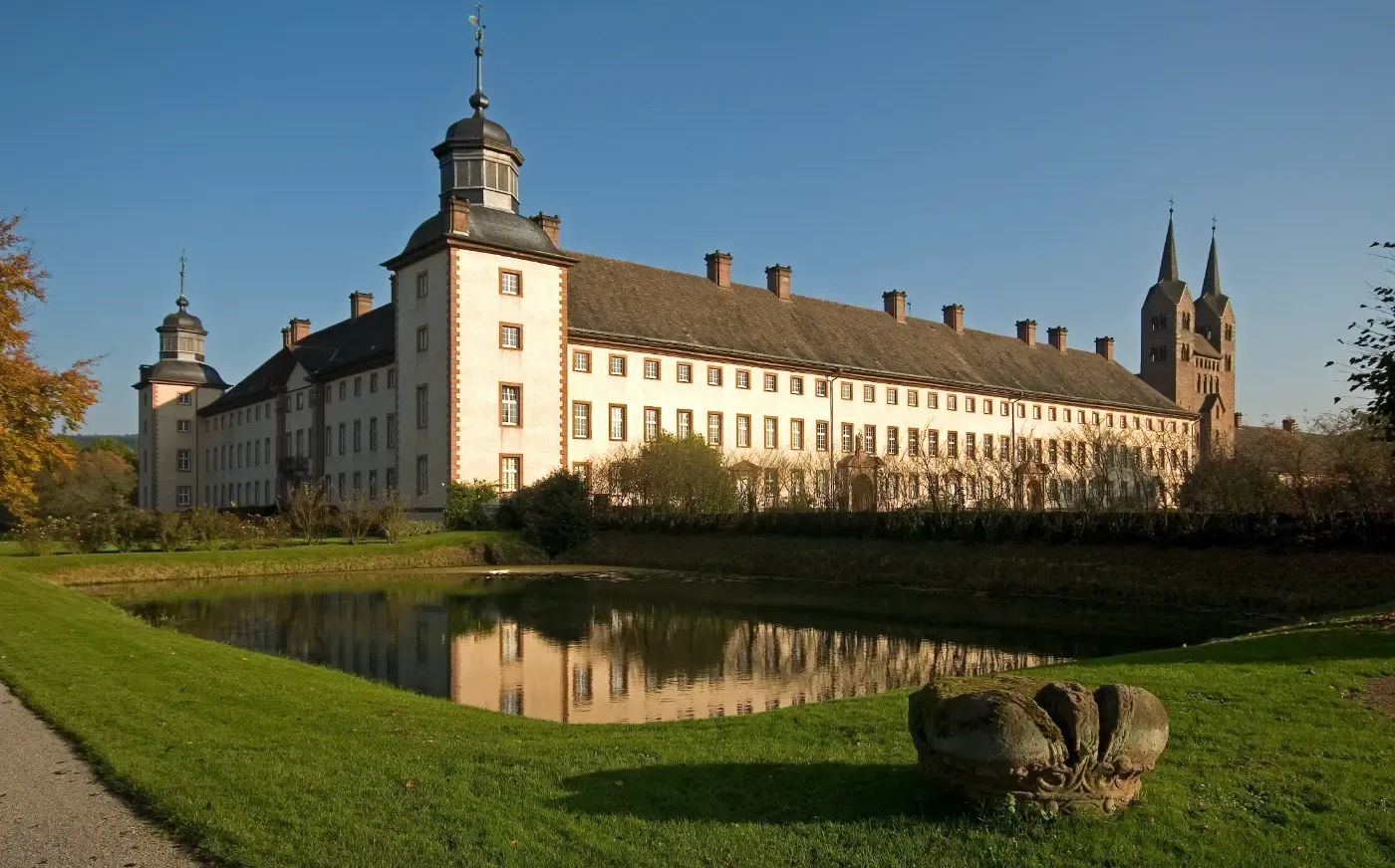 Ein langes, weißes, historisches Gebäude mit einem Turm neben einem spiegelnden Teich auf einer grasbewachsenen Rasenfläche unter klarem, blauem Himmel.