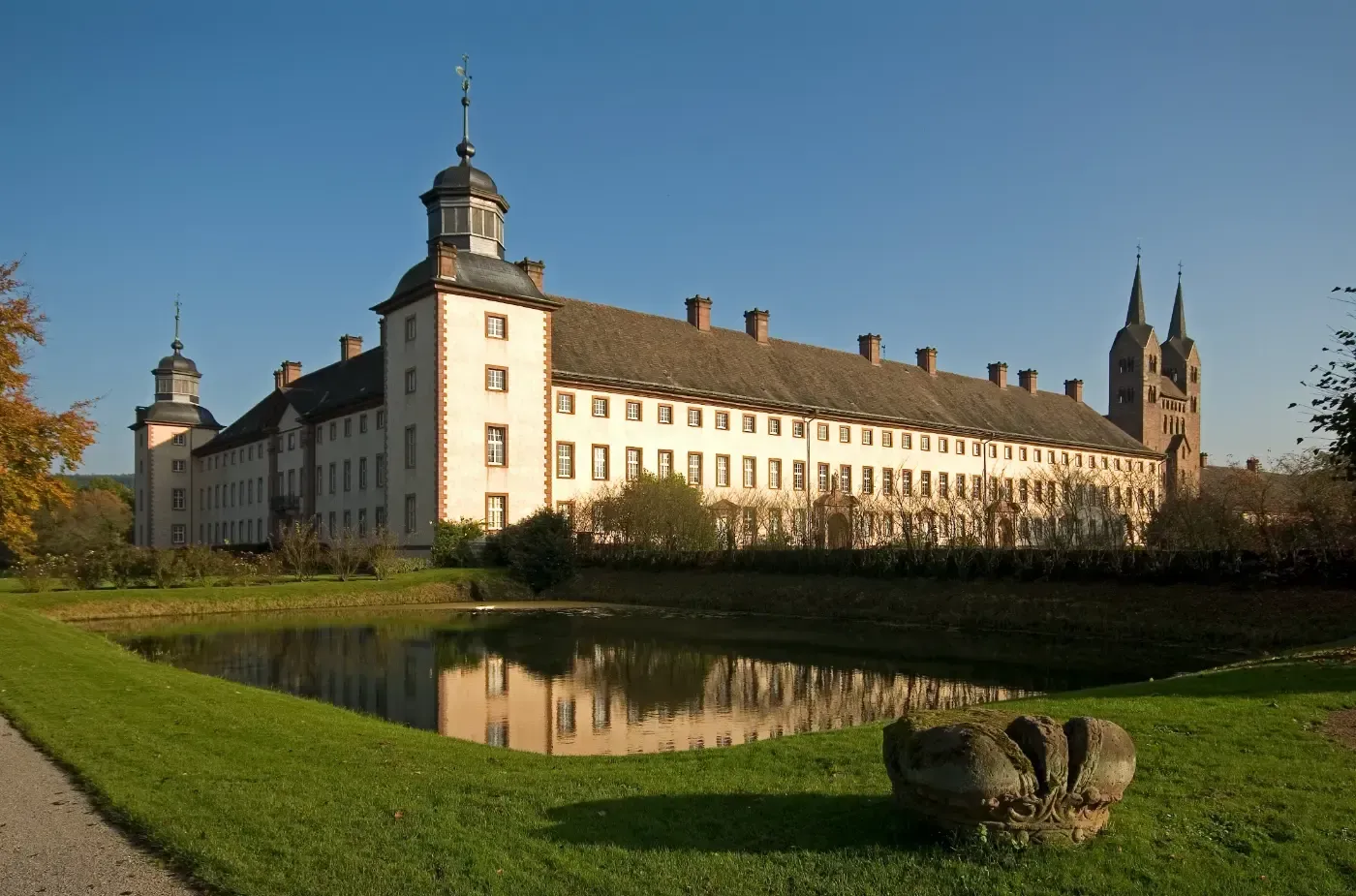 An einem sonnigen Tag überblickt ein langes, hellfarbiges historisches Gebäude mit Ecktürmen einen spiegelnden Teich.