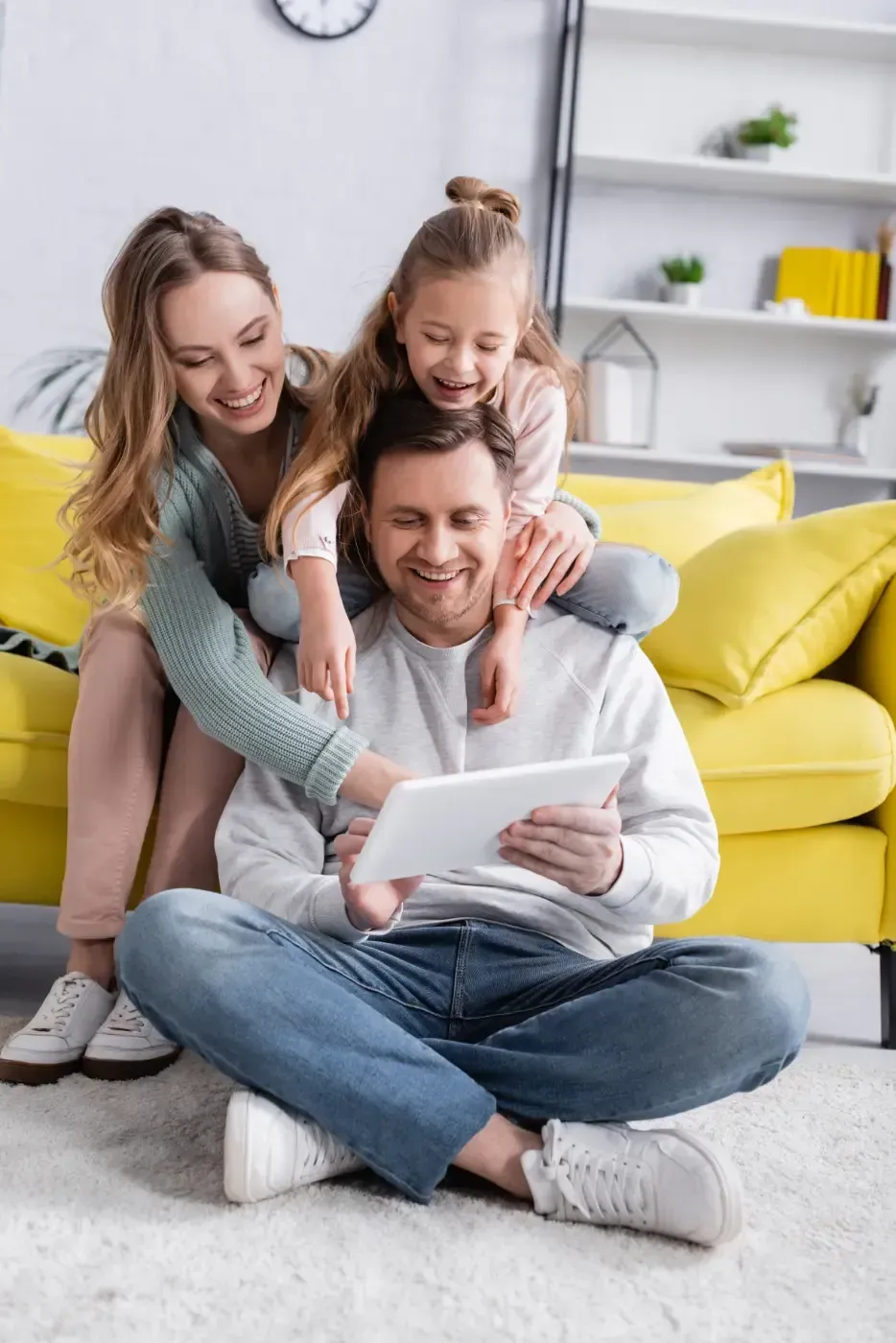 Eine Familie sitzt zusammen in einem Wohnzimmer und lächelt, während sie auf ein Tablet schaut, das von einer Person gehalten wird, die auf dem Boden sitzt.