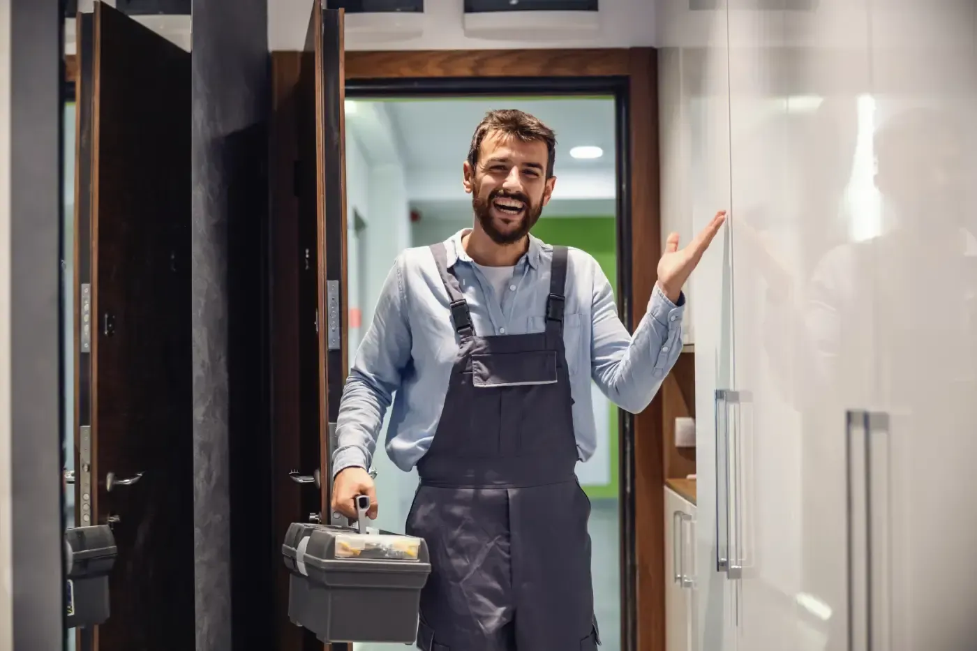 Ein lächelnder Fachmann in dunkler Arbeitskleidung und hellem Hemd hält einen Werkzeugkasten in der Hand und deutet auf weiße Schränke.