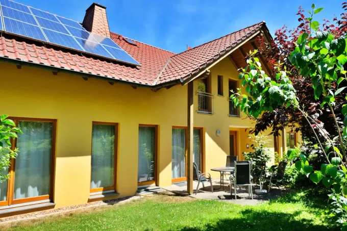 Ein gelbes zweistöckiges Haus mit rotem Ziegeldach, Solarpaneelen und einer Terrasse auf einem sonnigen Rasen.