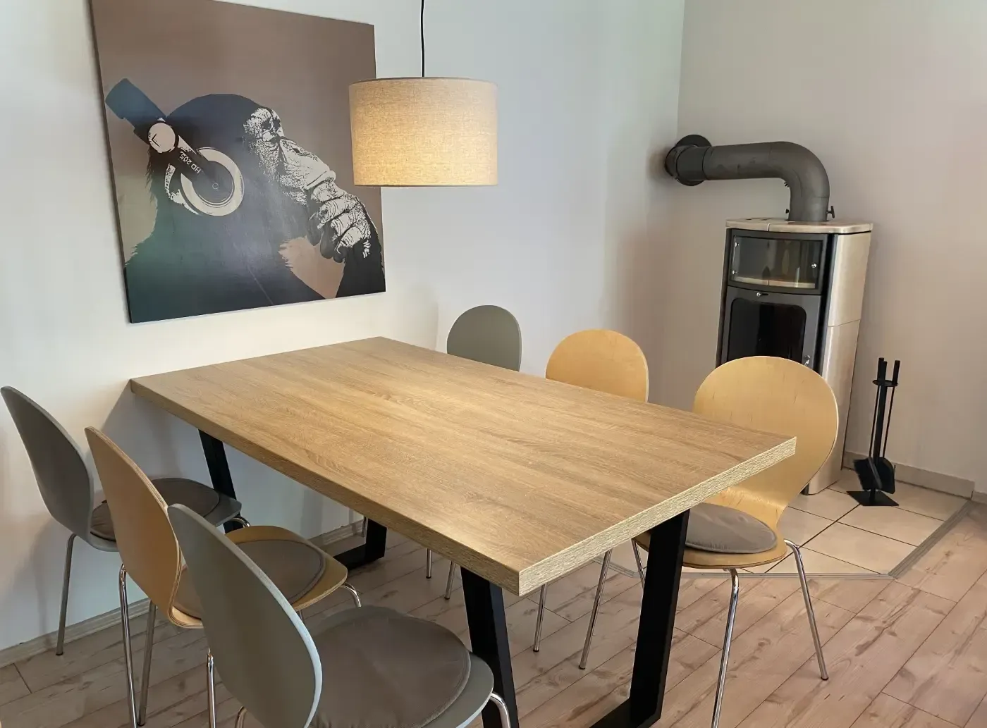 Esszimmer mit einem Holztisch, Stühlen in verschiedenen Grau- und Hellholztönen, einer Pendelleuchte und einem Holzofen.