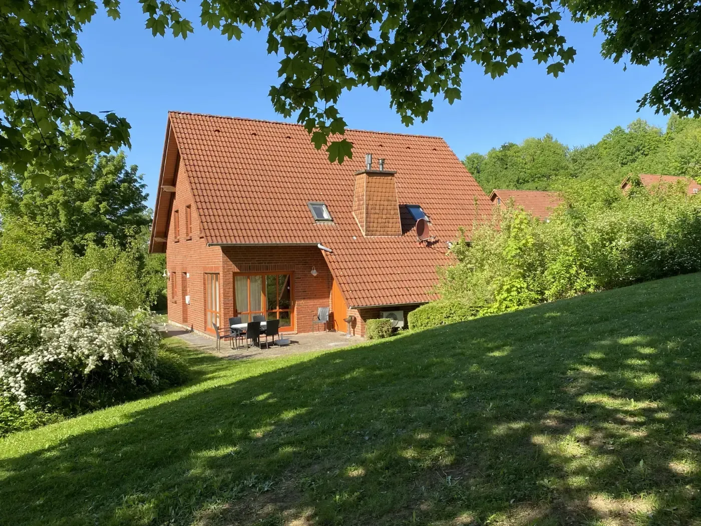 Ein Backsteinhaus mit rotem Ziegeldach und sonniger Terrasse, umgeben von einem üppigen grünen Rasen und Bäumen an einem klaren Tag.