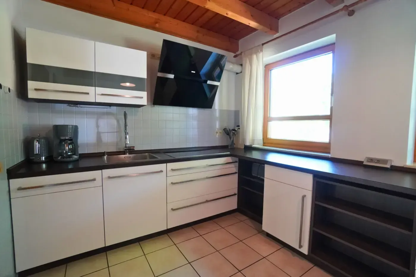 Eine Küche mit weißen Schränken, dunklen Arbeitsplatten, einer schwarzen Dunstabzugshaube, einem gefliesten Boden und einem Fenster mit Holzrahmen.