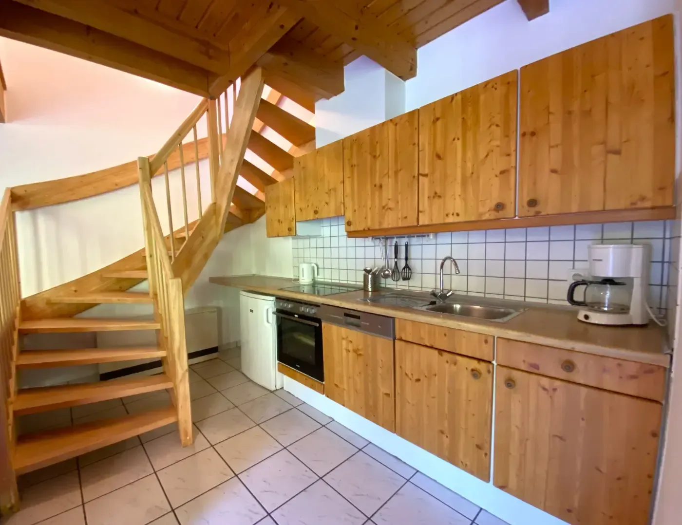 Eine Holzküche mit Fliesenboden, links eine Treppe und auf der Arbeitsfläche eine weiße Kaffeemaschine.