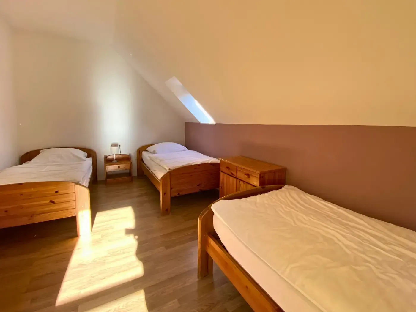 Drei hölzerne Einzelbetten in einem sonnendurchfluteten Dachgeschosszimmer mit beigen Wänden und hellem Holzboden.