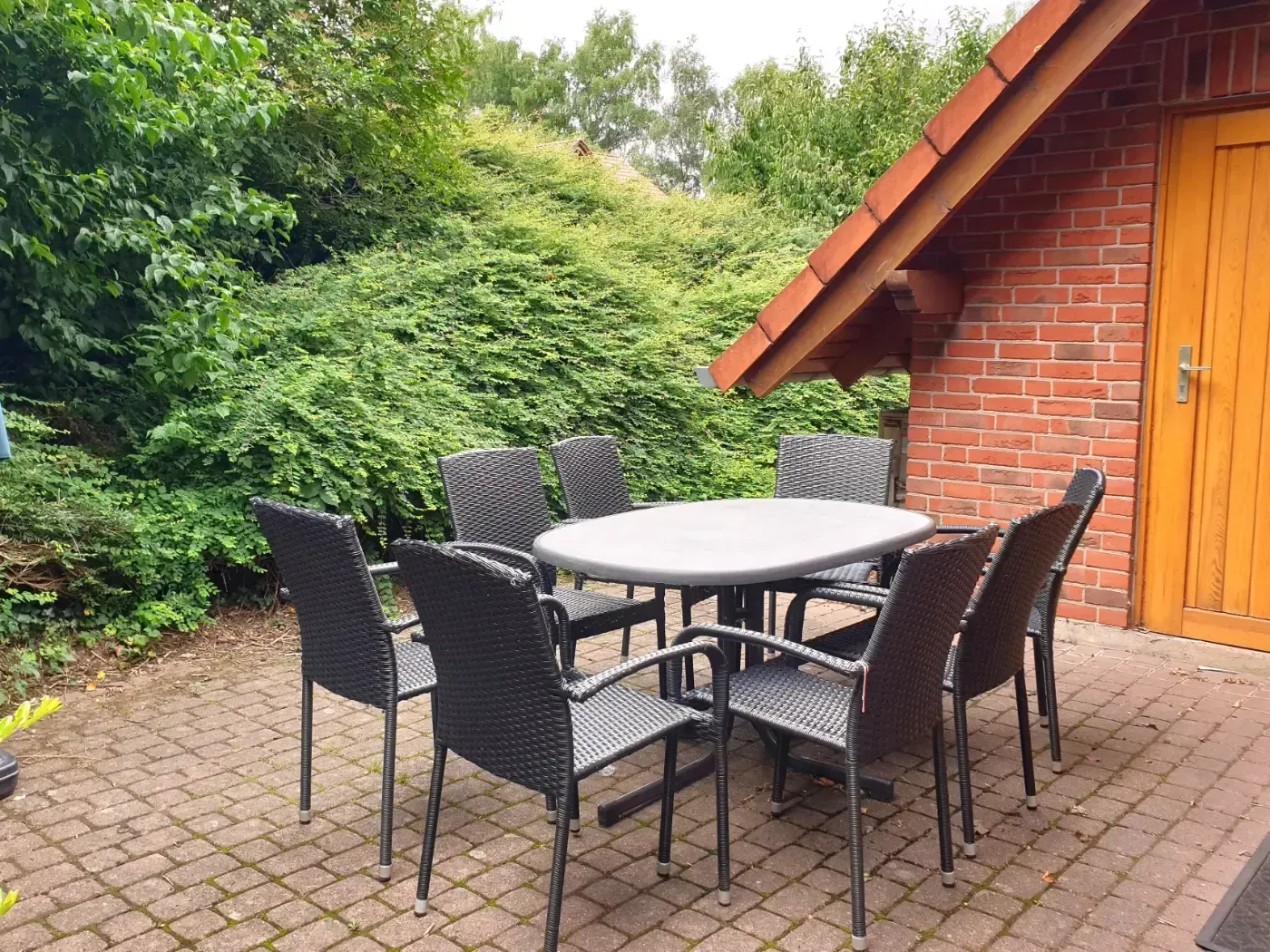 Eine Terrasse mit einem hellgrauen ovalen Tisch und sechs schwarzen Korbstühlen auf einer Ziegelfläche neben einem Backsteingebäude.