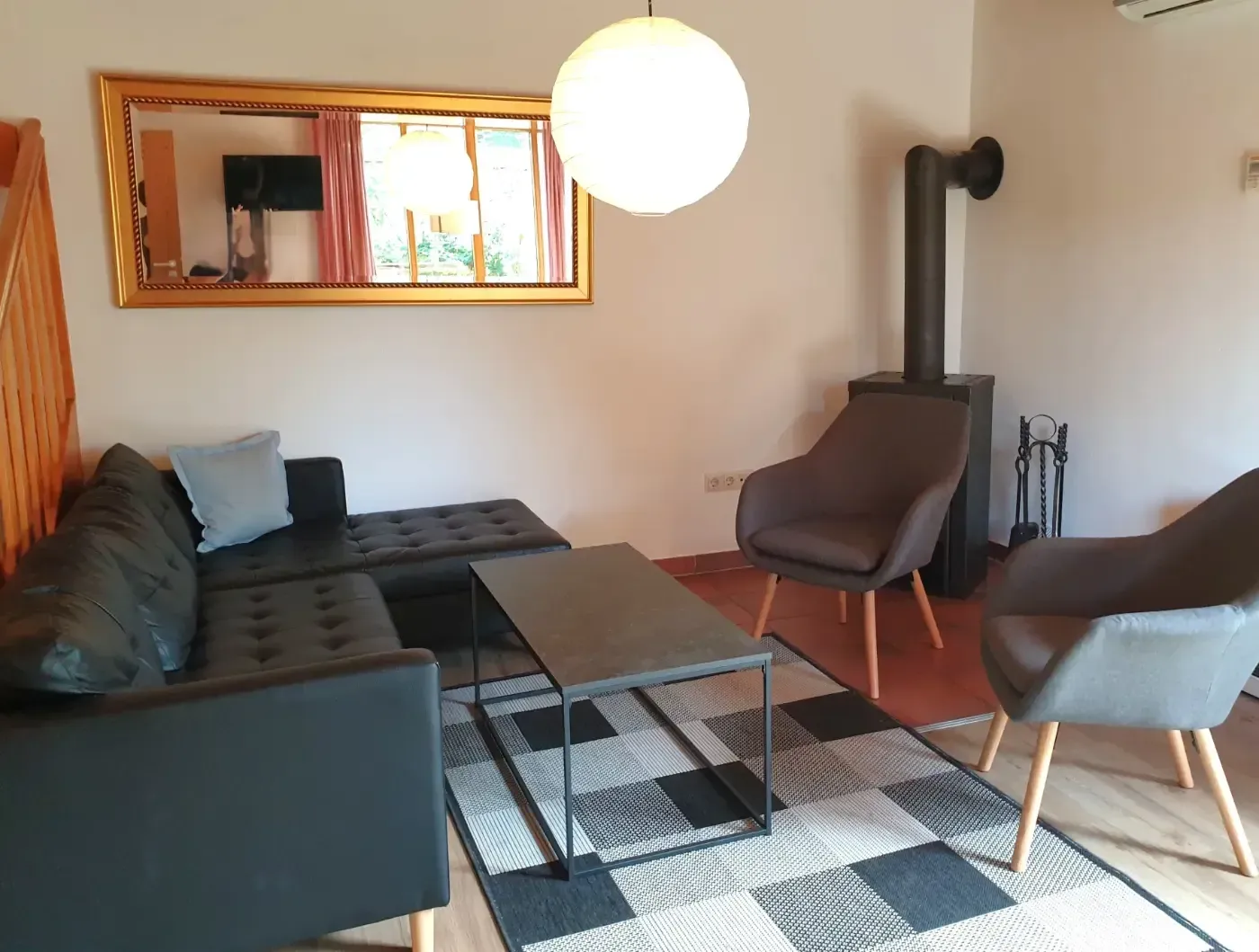 Ein Wohnzimmer mit einer dunklen Ledersitzgruppe, zwei grauen Sesseln, einem Couchtisch, einem Holzofen und einem Wandspiegel.