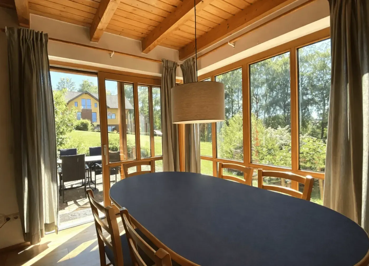 Ein Esszimmer mit einem dunklen ovalen Tisch, Holzstühlen und einer Hängelampe, das durch große Fenster auf einen Garten blickt.