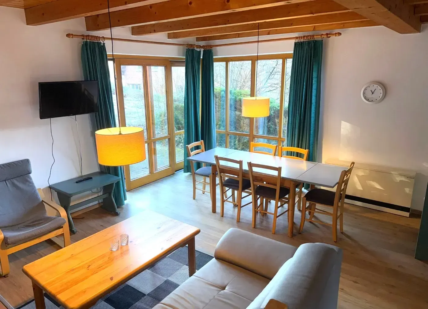 Ein Wohn- und Essbereich mit Sofa, hölzernem Couchtisch, Essgruppe, Fernseher und großen Fenstern mit türkisfarbenen Vorhängen.