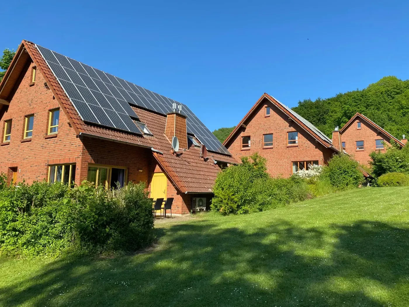 Drei rote Backsteinhäuser mit Satteldächern, eines davon mit Solarpaneelen, stehen auf einem grasbewachsenen Hügel unter klarem, blauem Himmel.