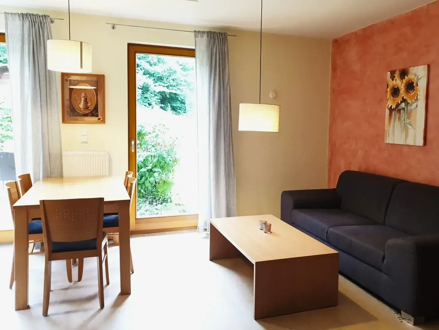 Ein Wohn- und Essbereich mit einem dunklen Sofa, einem hellen Couchtisch und einer Essgruppe aus Holz sowie einem Wandbild mit Sonnenblumenmotiv.