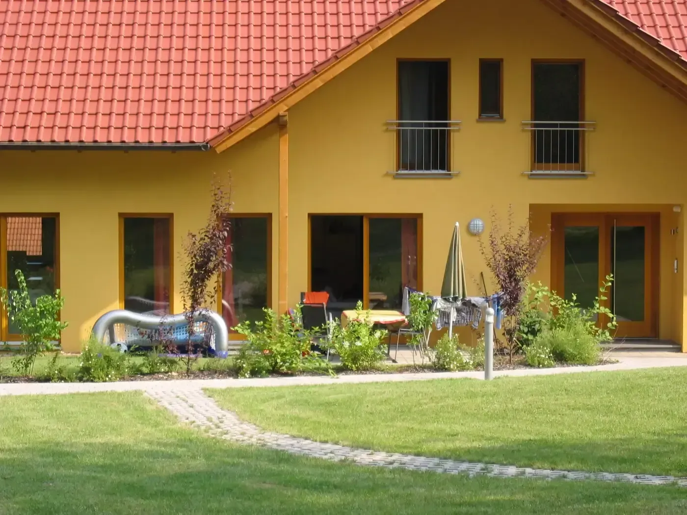 Gelbes Haus mit rotem Ziegeldach, Terrasse und einem Steinweg, der über einen grünen Rasen führt.