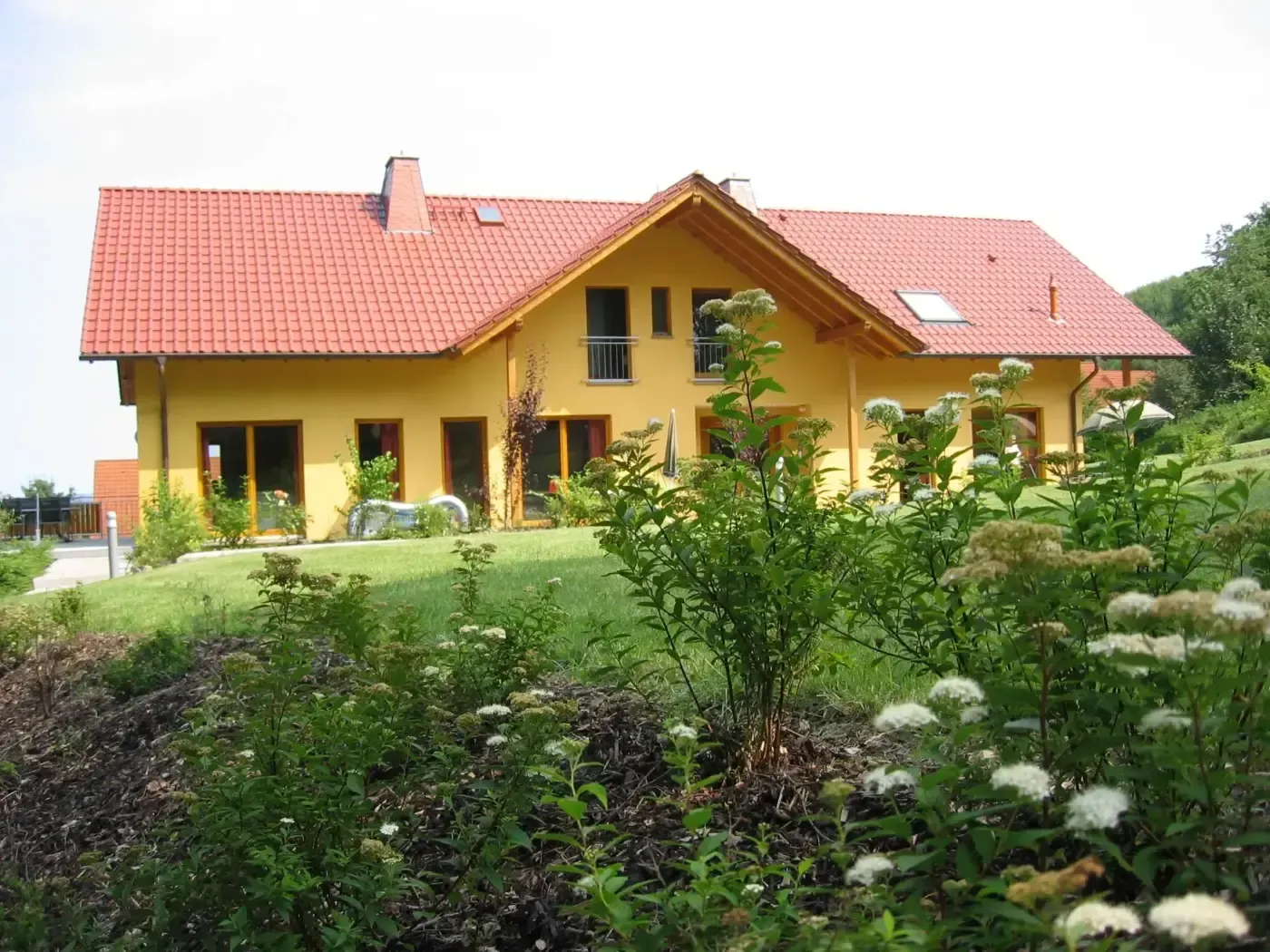 Hinter einem Garten mit grünen Pflanzen und weiß blühenden Sträuchern steht ein zweistöckiges gelbes Haus mit rotem Ziegeldach.