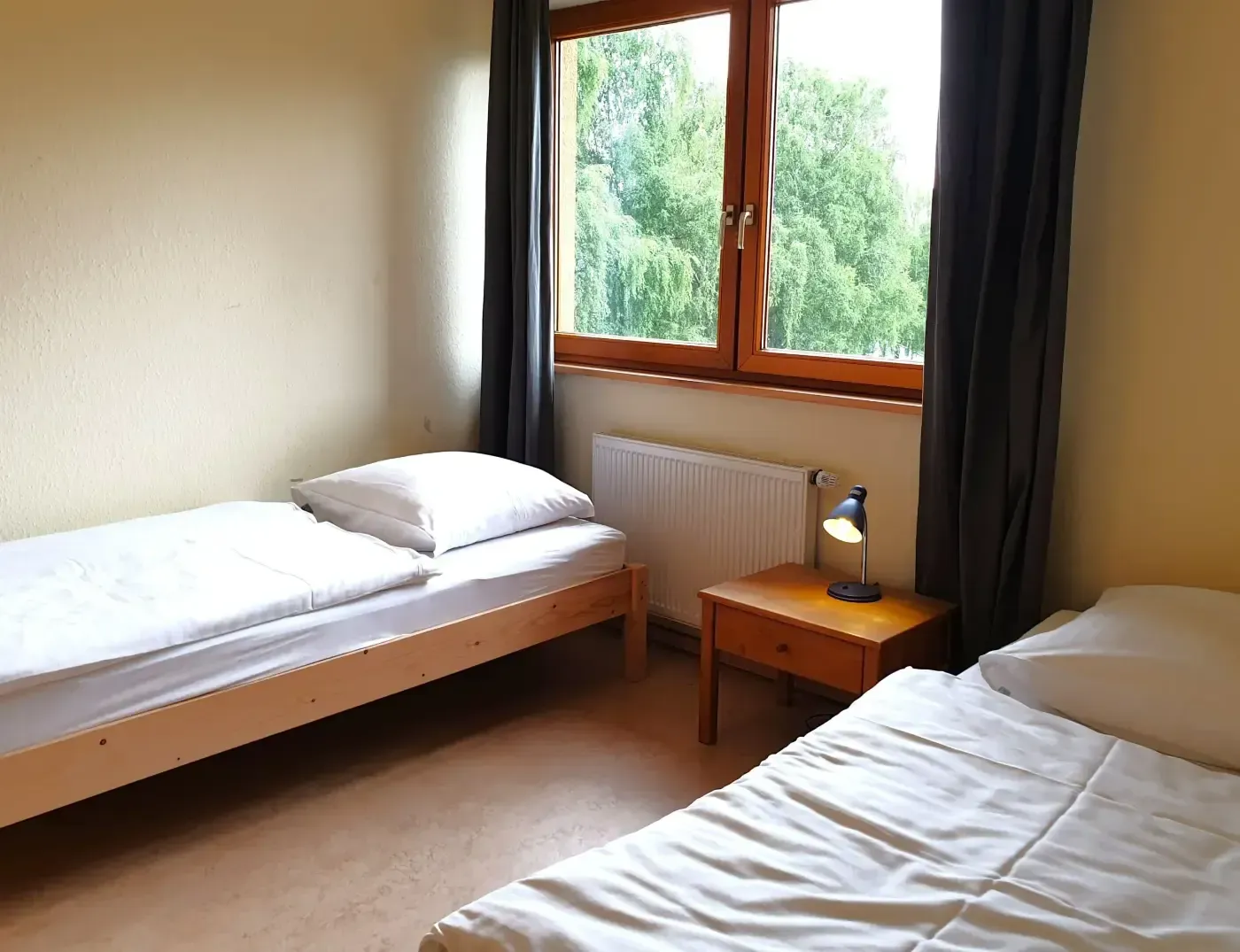 Ein Schlafzimmer mit zwei Einzelbetten, einem hölzernen Nachttisch mit Lampe und einem Fenster mit grauen Vorhängen und einem Heizkörper darunter.