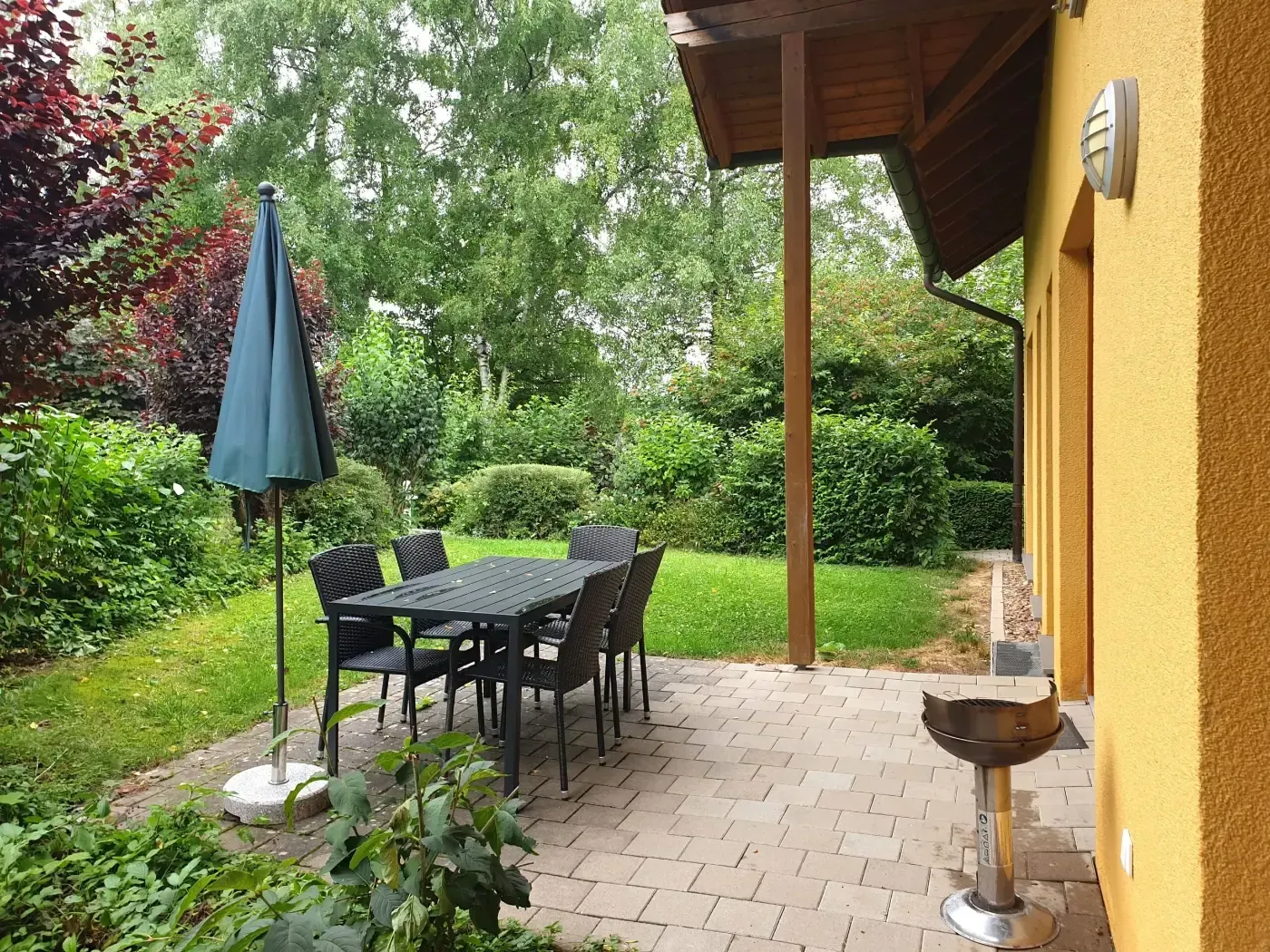 Eine Terrasse mit einem dunklen Esstisch und Stühlen unter einem Sonnenschirm, neben einem gelben Haus und einem kleinen Grill in einem Garten.
