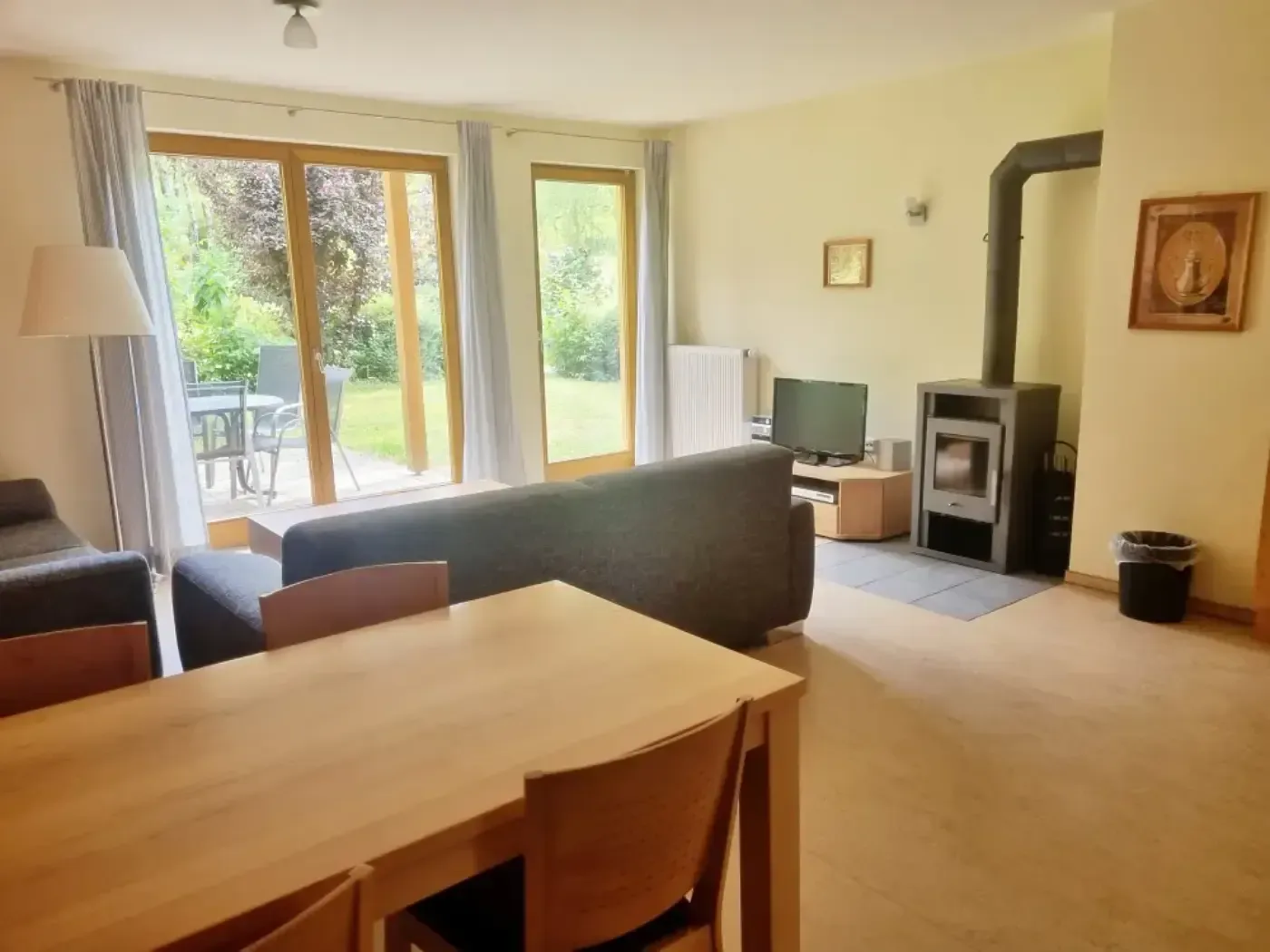 Ein helles Wohnzimmer mit einem Esstisch im Vordergrund, einem grauen Sofa, einem Holzofen und Glastüren zur Terrasse.