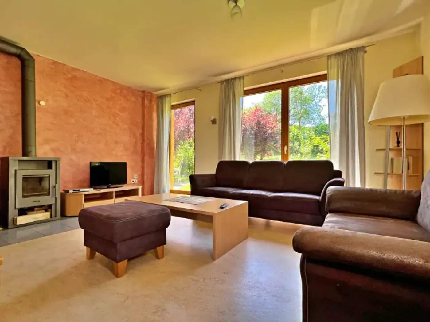 Ein Wohnzimmer mit braunem Sofa und Hocker, einem Kamin an einer strukturierten pfirsichfarbenen Wand und großen Fenstern mit Blick nach draußen.
