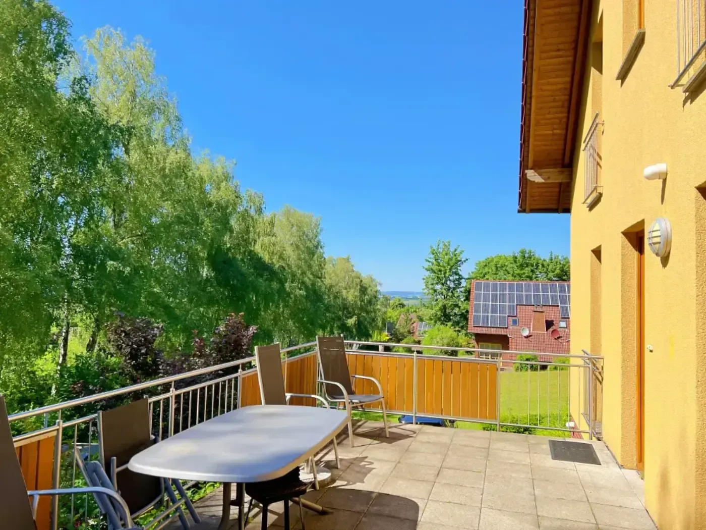 Ein sonniger Balkon mit Gartentisch und Stühlen, mit Blick auf grüne Bäume und ein Haus mit Solaranlage in der Ferne.