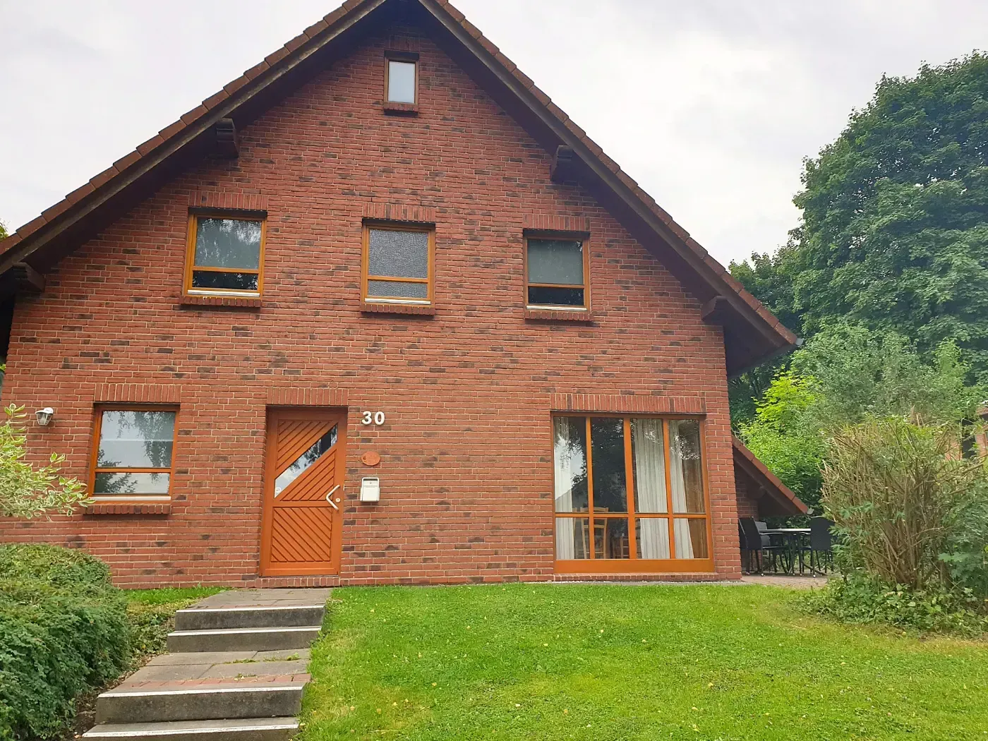 Ein zweistöckiges Haus aus roten Backsteinen mit Holzverzierungen, einer Eingangstür und einem grünen Rasen mit einer Steintreppe im Vordergrund.