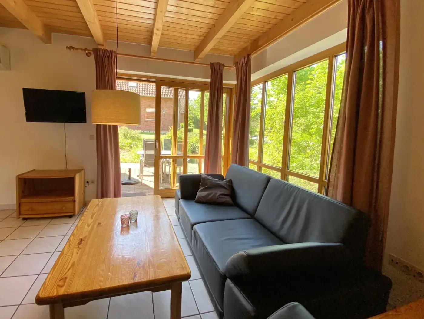 Ein gemütliches Wohnzimmer mit einem dunklen Sofa, einem Couchtisch aus Holz, einem an der Wand montierten Fernseher und großen Fenstern mit Blick auf die Bäume.