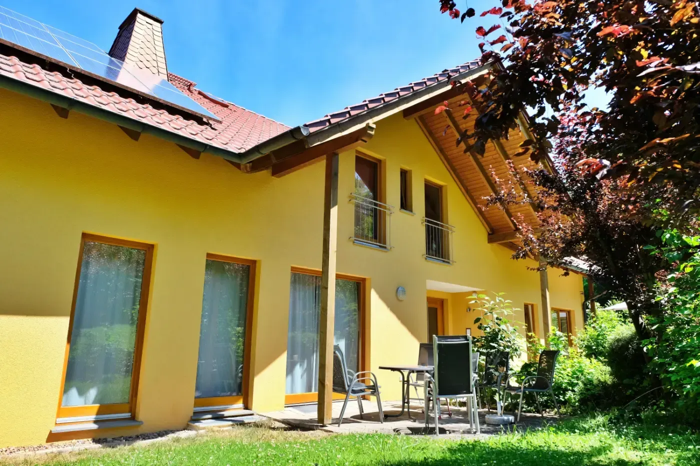 Ein gelbes zweistöckiges Haus mit rotem Ziegeldach, großen Glastüren und einer Terrasse auf einem sonnigen, grünen Rasen.