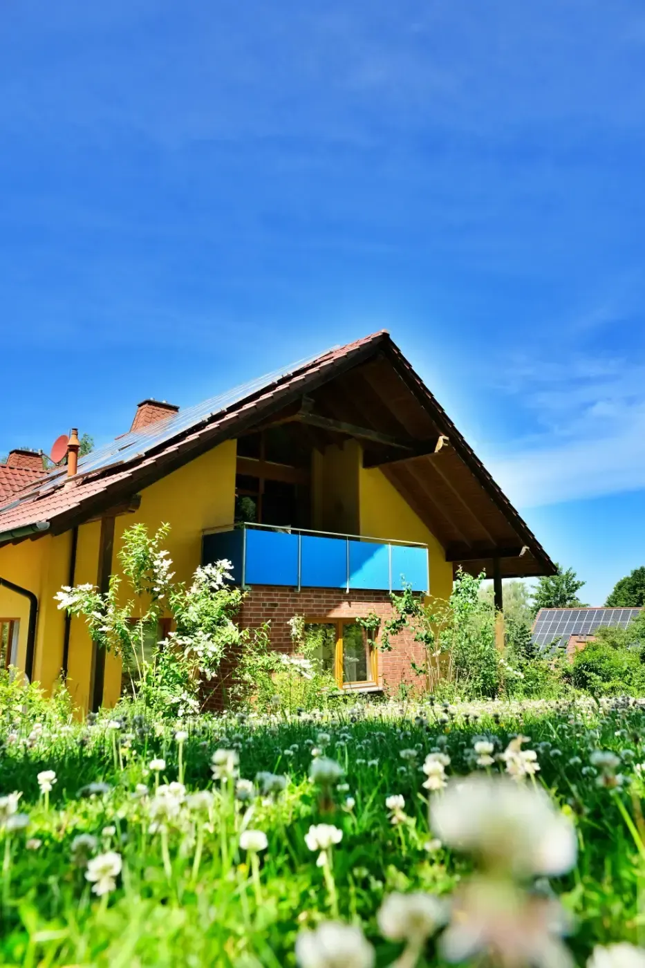Hinter einer Wiese mit weißen Kleeblüten unter blauem Himmel steht ein gelbes Haus mit Balkon und braunem Dach.