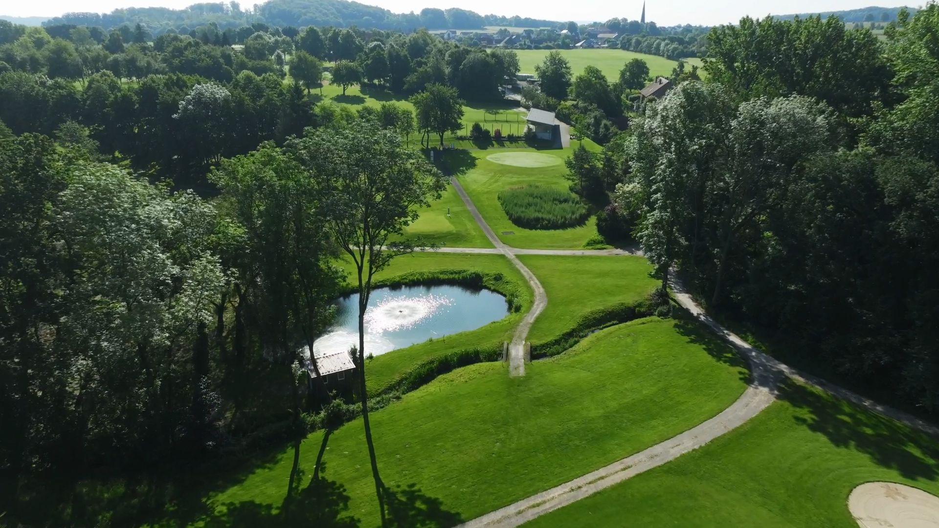 Luftaufnahme eines üppig grünen Golfplatzes mit einem kleinen Teich mit Springbrunnen, gewundenen Pfaden und umliegenden Bäumen.