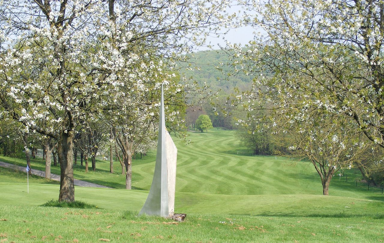 Auf dem grünen, gemähten Fairway eines Golfplatzes steht unter klarem Himmel eine weiße, abstrakte Skulptur, umgeben von blühenden Bäumen.
