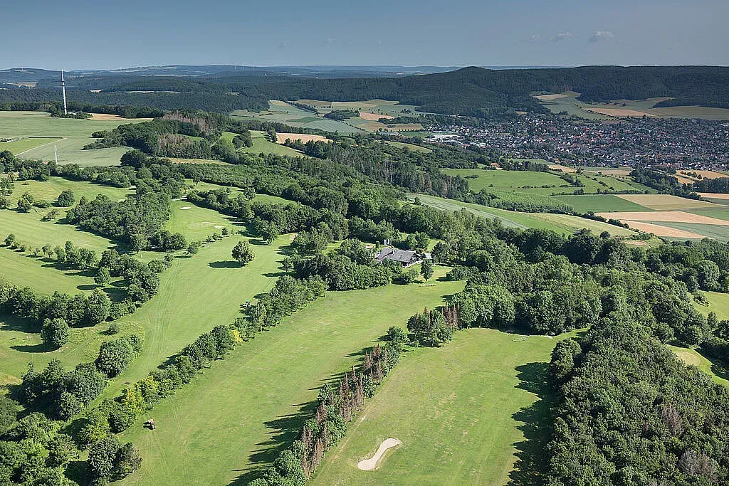 Luftaufnahme einer grünen Landschaft mit Golfplatz, vereinzelten Bäumen und einer Stadt in der Ferne unter klarem, blauem Himmel.