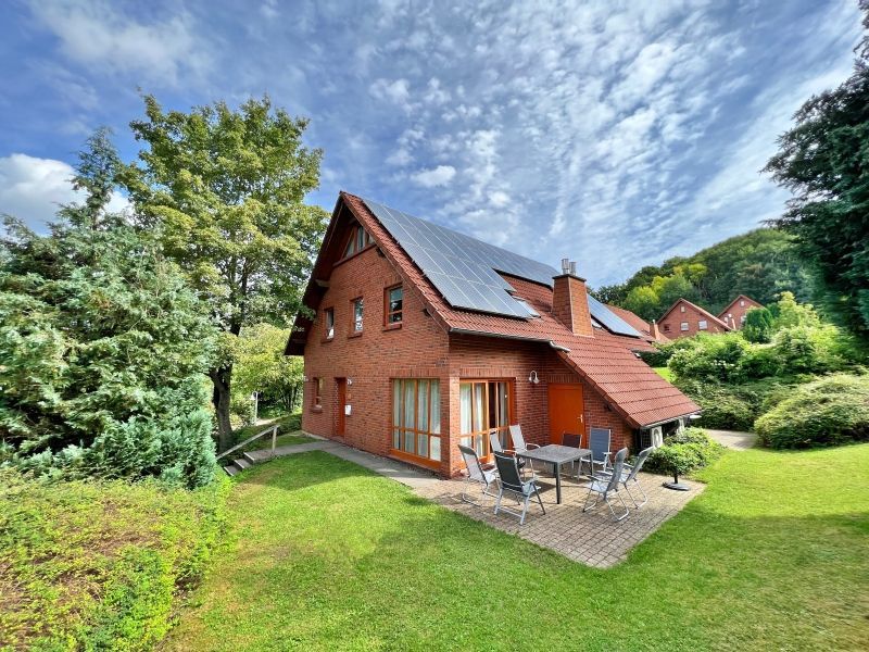 Ein Backsteinhaus mit dunklem Dach und Solarpaneelen, mit einer Terrasse und Gartenmöbeln inmitten eines grünen Gartens.