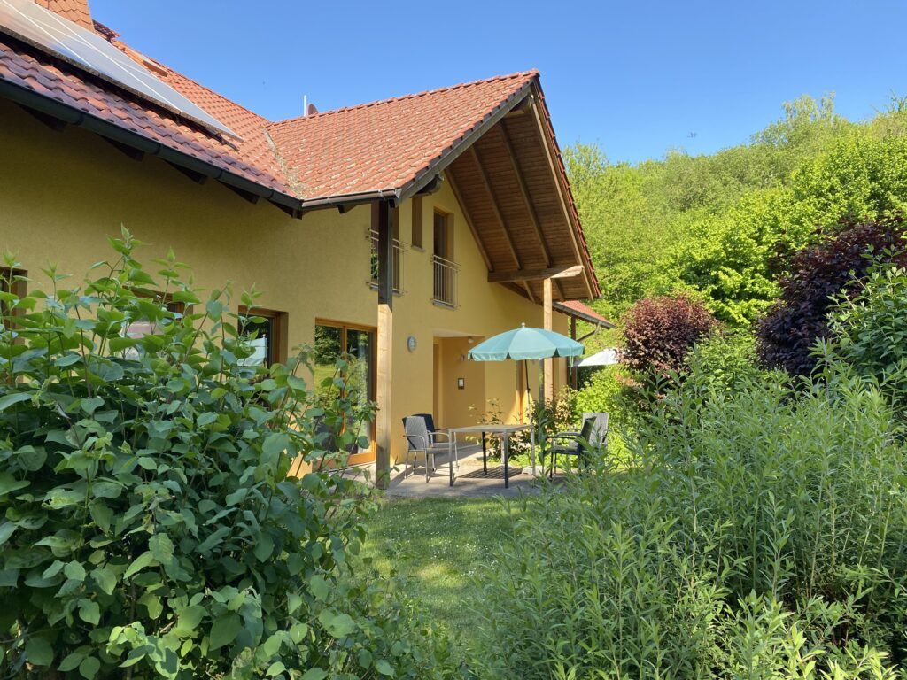 Ein cremefarbenes Haus mit Ziegeldach und Terrasse, umgeben von üppigem Grün im Garten unter klarem, blauem Himmel.
