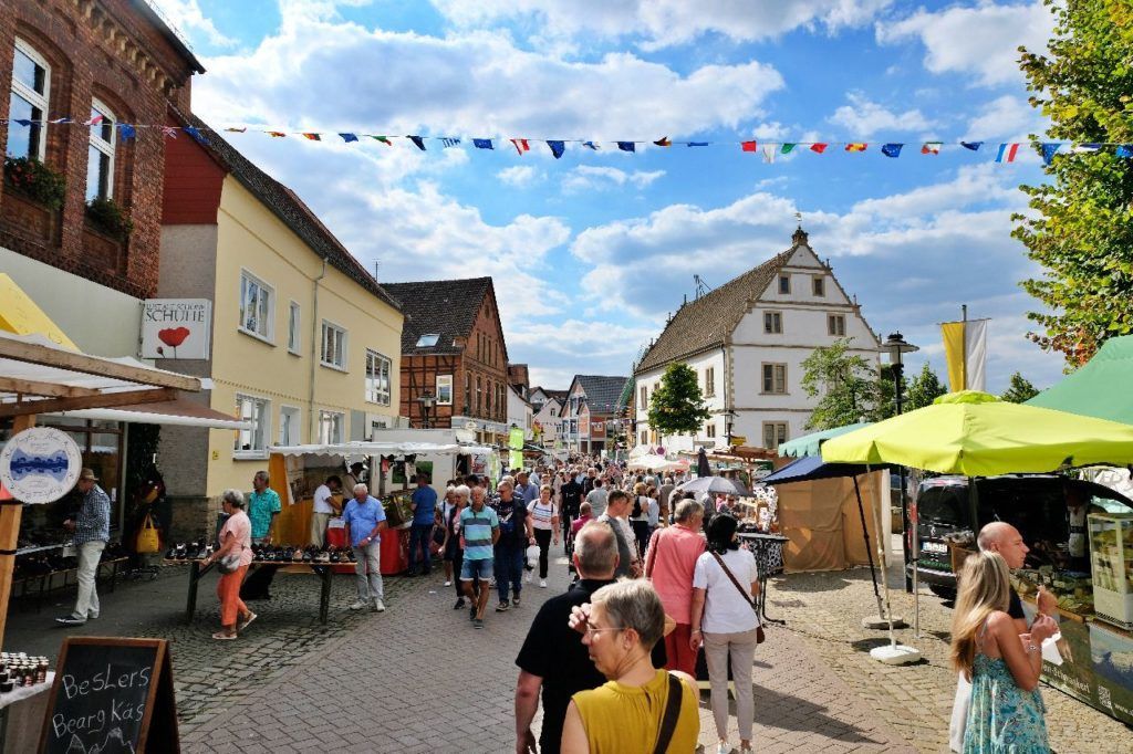 Ein geschäftiger Straßenmarkt auf einer Kopfsteinpflasterstraße, gesäumt von historischen Gebäuden, farbenfrohen Ständen und festlicher Wimpelkette.